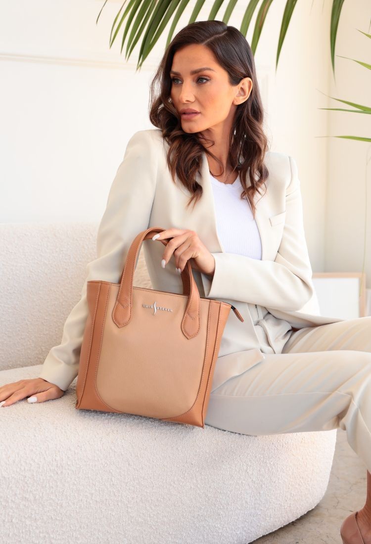 Luis Steven small bucket tote in tan pebble leather held by a woman in a cream blazer sitting on a white sofa.